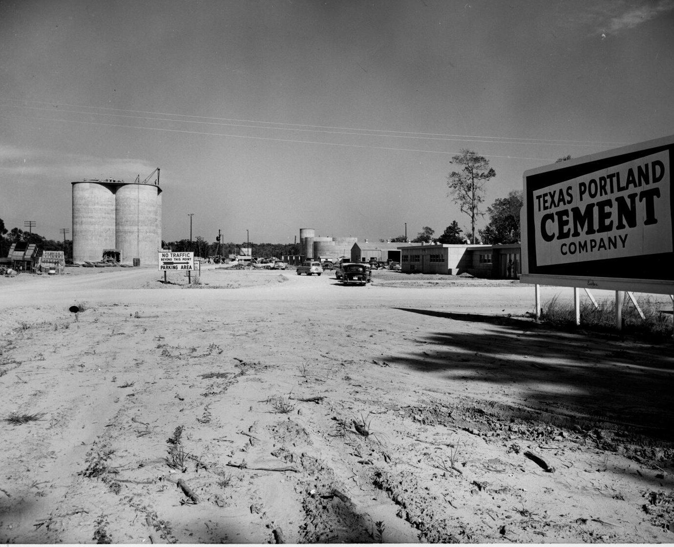 A bare, sun-bleached lot in Cement City, West Dallas: twin concrete silos at left, a construction shed and low office buildings at center, and a roadside sign reading TEXAS PORTLAND CEMENT COMPANY at right, undated.